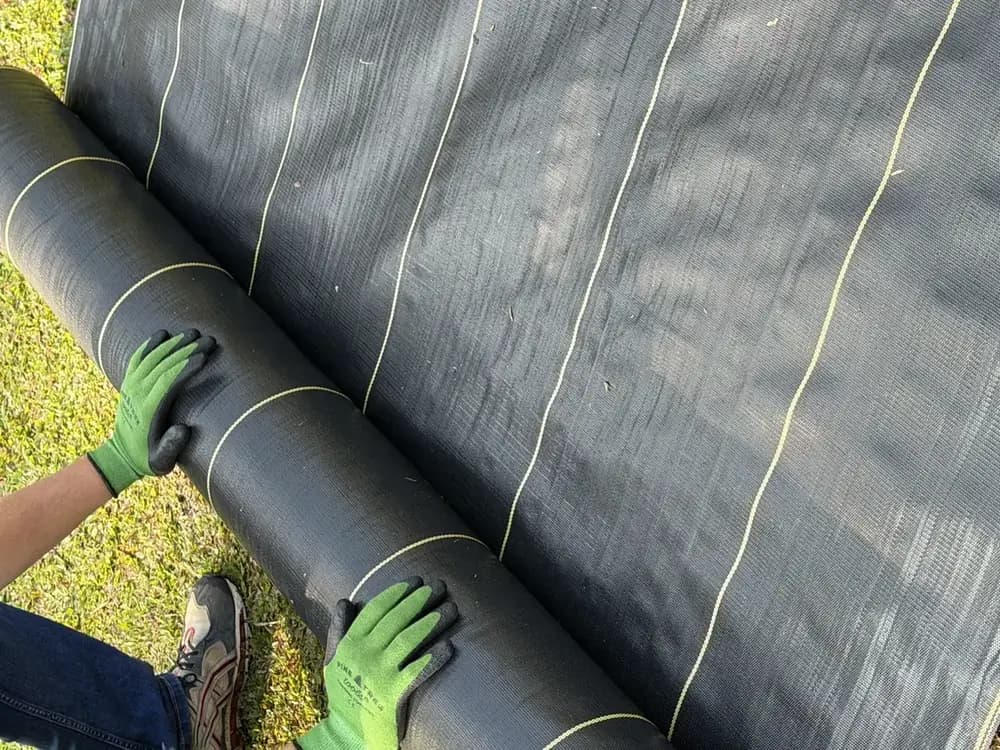 Installer handling a roll of DOTDAY SHIELD 3.2 oz woven weed barrier fabric over grass