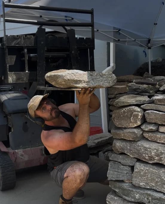 Jeff lifting a stone slab overhead beside a stacked stone wall