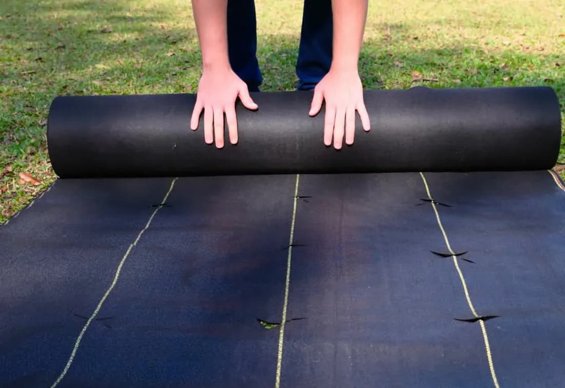 Heavy duty landscape fabric: hands unrolling a DOTDAY XBAR 5oz dual-layer woven roll with yellow guide lines across a lawn