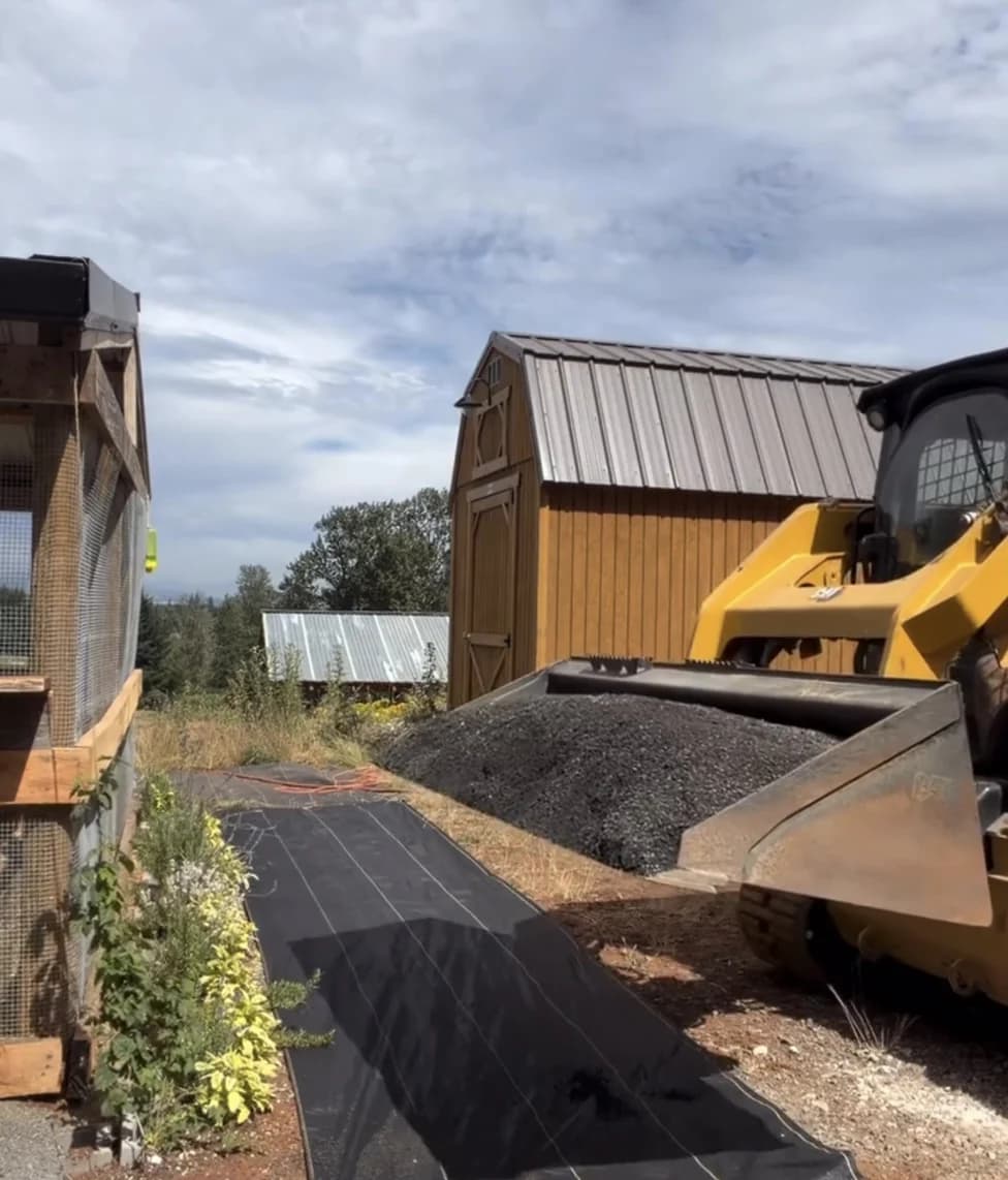 A long run of DOTDAY XBAR 5oz landscape fabric rolled out with yellow guide lines as a skid-steer brings gravel to spread over it