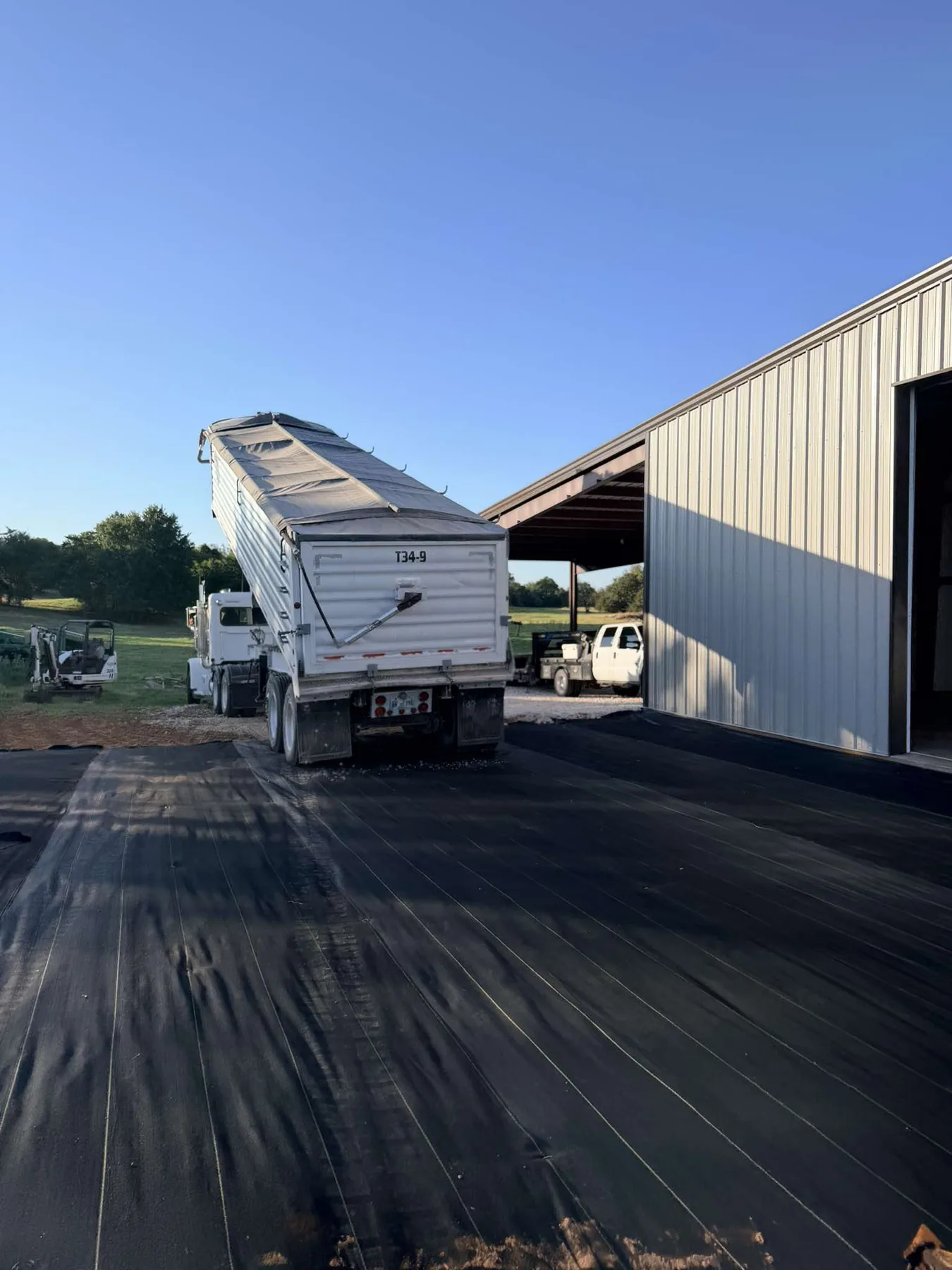Loaded end dump trailer backed onto bare DOTDAY XBAR fabric in front of the shop before tipping gravel