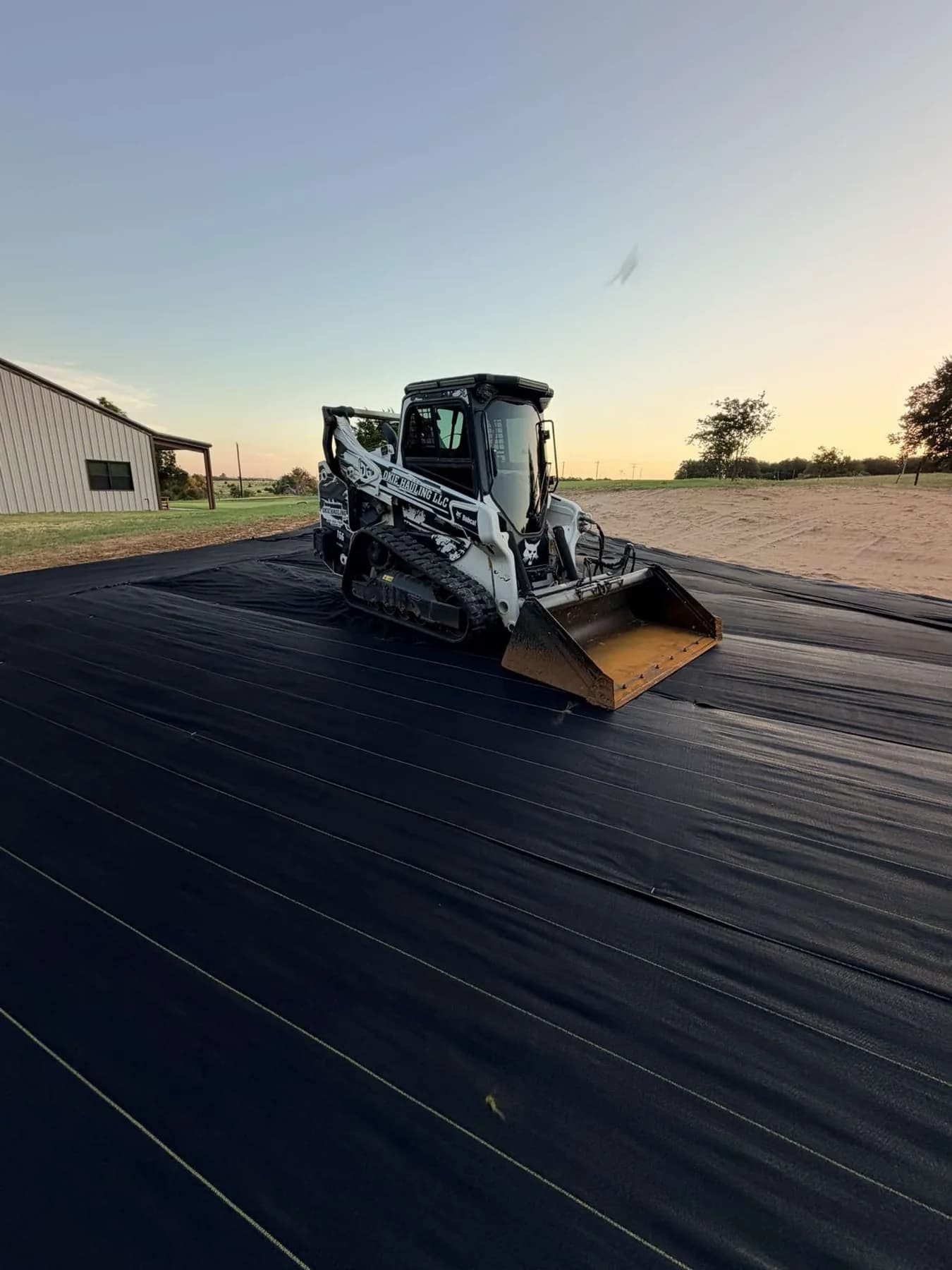 Okie Hauling skid steer parked on DOTDAY XBAR 5oz fabric across a shop pad at sunset near Ringwood, Oklahoma