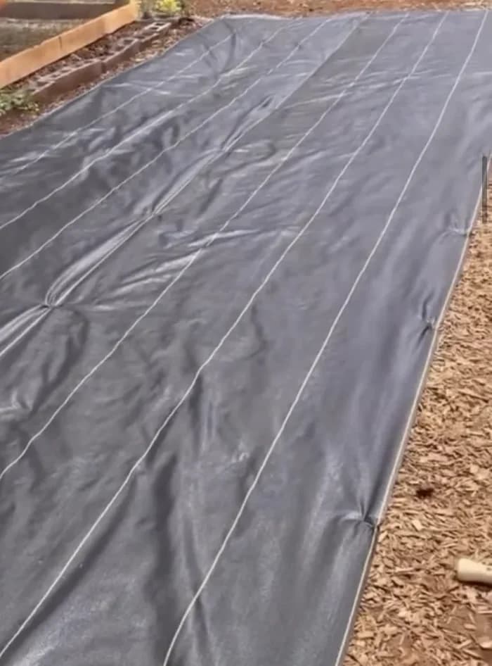 Black woven fabric with guide lines laid flat over a garden bed beside wood chips and raised beds