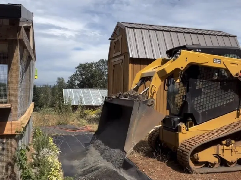 Yellow skid steer dumping gravel from its bucket onto black XBAR fabric beside a chicken coop run and a wooden shed