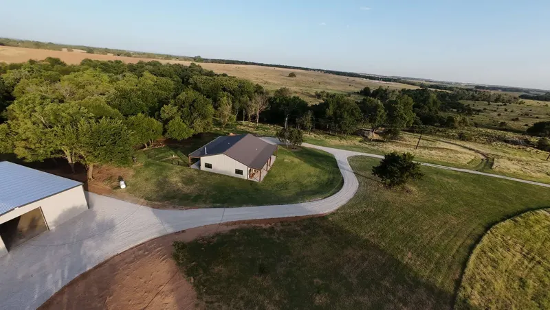 Drone view of a finished white gravel driveway curving from a metal shop past a house and lawn, built over DOTDAY XBAR