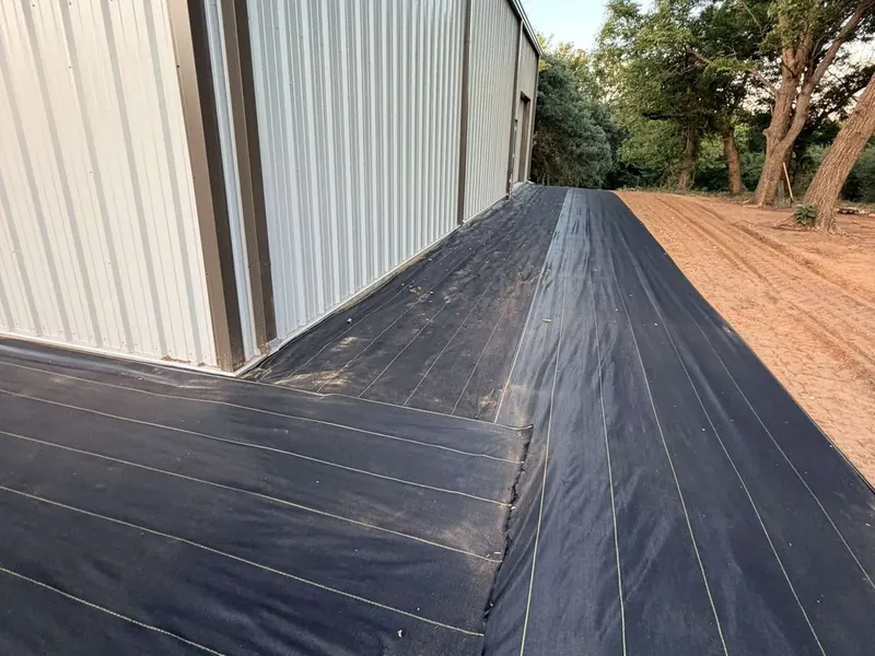 Overlapped runs of black XBAR fabric with yellow guide lines along a white metal shop wall before gravel