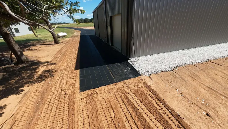 Black XBAR fabric rolled along the side of a metal shop on graded red dirt, with white gravel already spread at the far end
